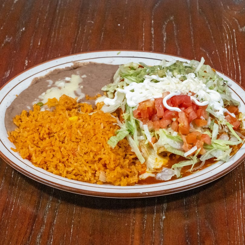 A plate of Mexican food with rice, refried beans, and enchiladas topped with lettuce, tomatoes, and sour cream.