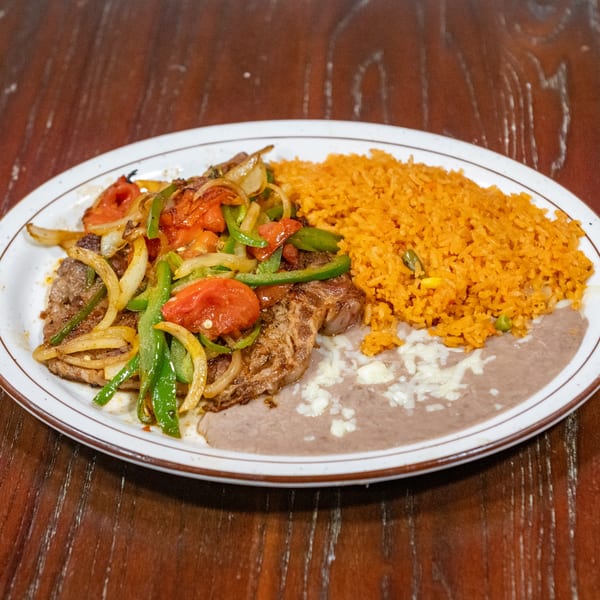 Steak with grilled onions, bell peppers, tomatoes, rice, and refried beans on a white plate.