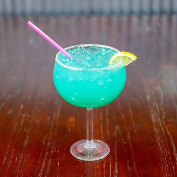 Bright blue cocktail with ice, a pink straw, and a lemon slice on a wooden table.
