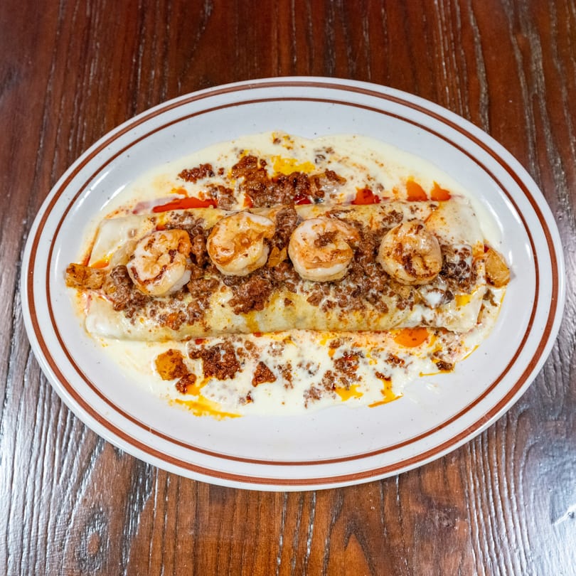 A white oval plate with a burrito covered in white sauce, ground meat, and shrimp.