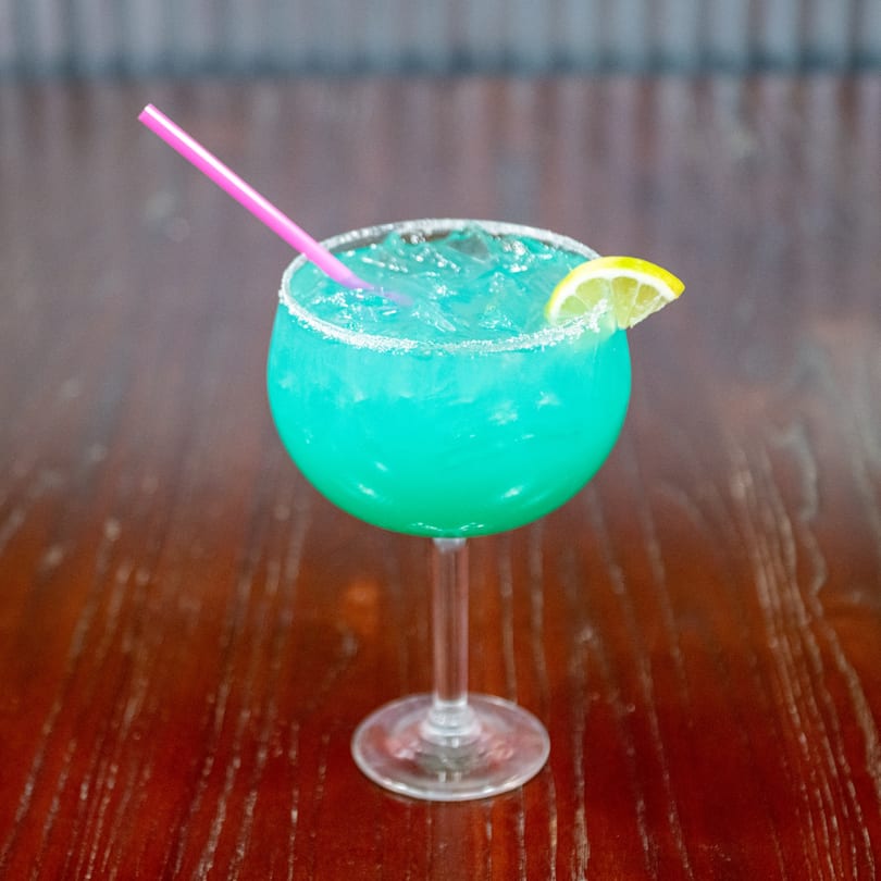 Bright blue margarita with a salted rim, lemon slice, and pink straw on a wooden table.