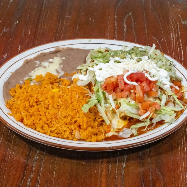 A plate of Mexican food with rice, refried beans, and enchiladas topped with lettuce, tomatoes, and sour cream.