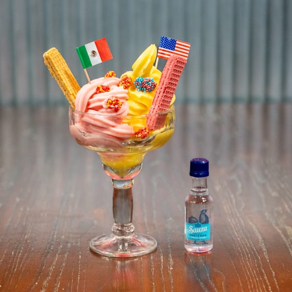 Pink and yellow soft serve in a glass with Mexican and American flags, next to a tequila bottle.