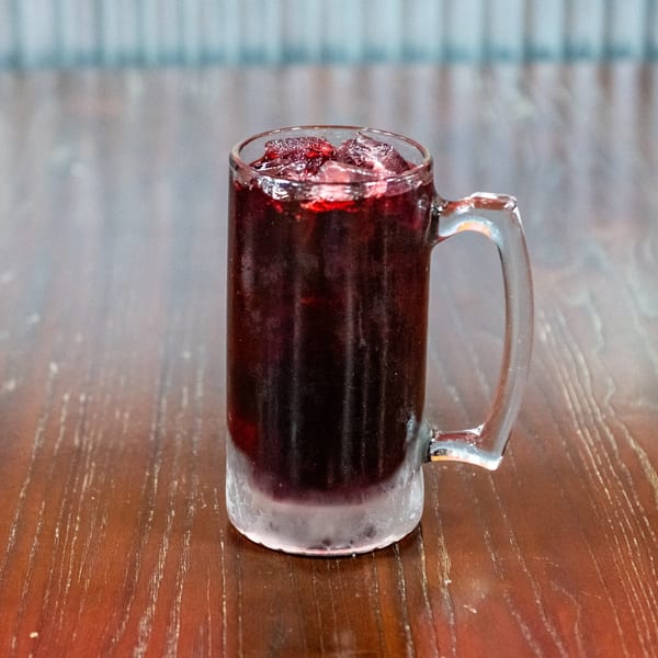 A frosty mug of dark red beverage with ice and fruit pieces sits on a wooden table.