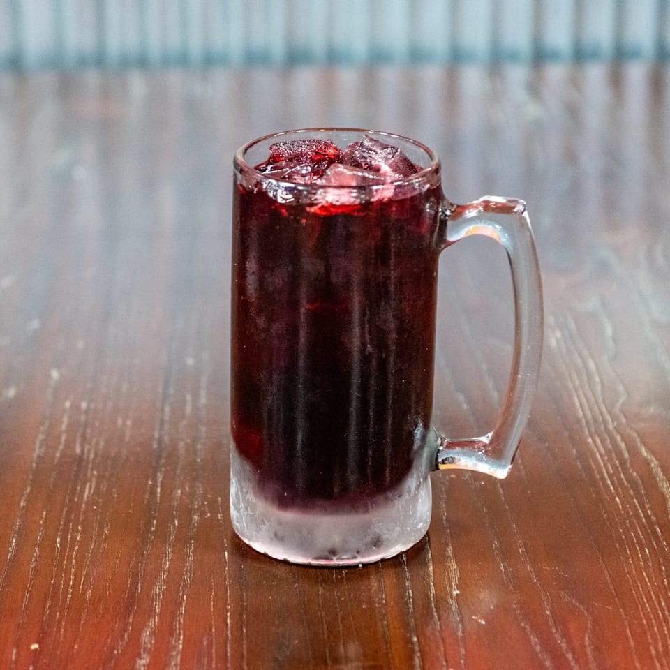 A frosty mug of dark red beverage with ice and fruit pieces sits on a wooden table.