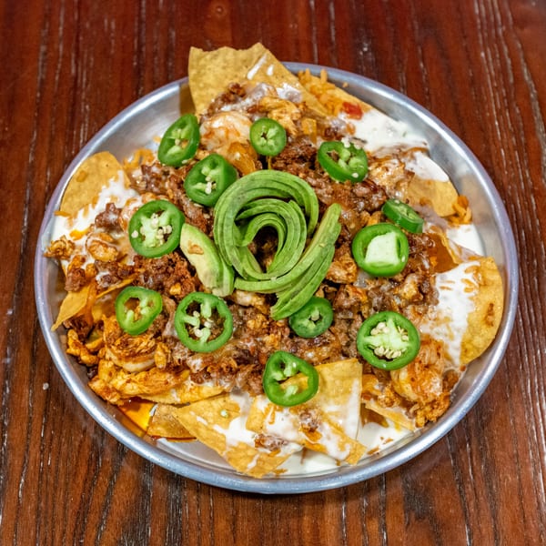 Nachos with ground meat, shrimp, cheese sauce, sliced jalapeños, and avocado on a metal plate.