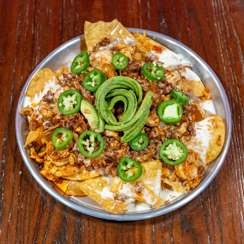 Nachos with ground meat, shrimp, cheese sauce, sliced jalapeños, and avocado on a metal plate.