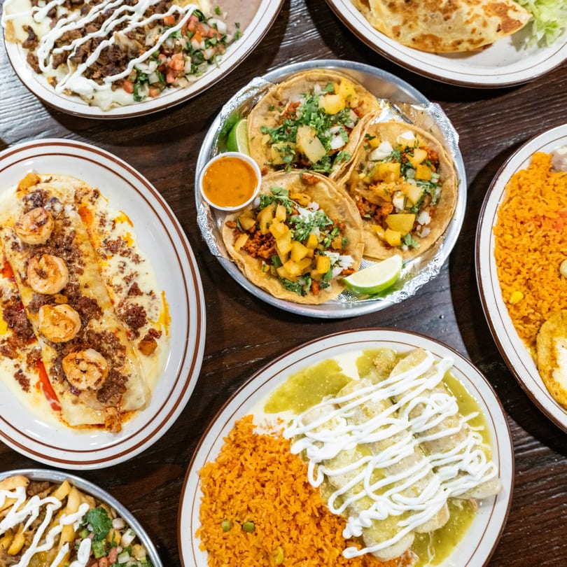 A top-down view of a wooden table laden with various Mexican dishes, including tacos, enchiladas, and rice.