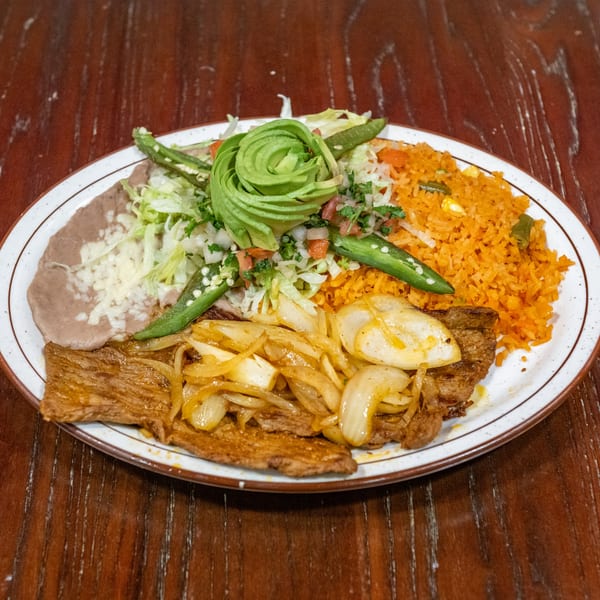 Carne asada with grilled onions, refried beans, rice, and a salad with avocado on a white plate.