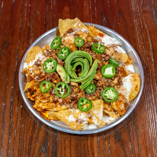 Loaded nachos with meat, cheese sauce, sliced jalapeños, and a swirl of avocado on a metal plate.