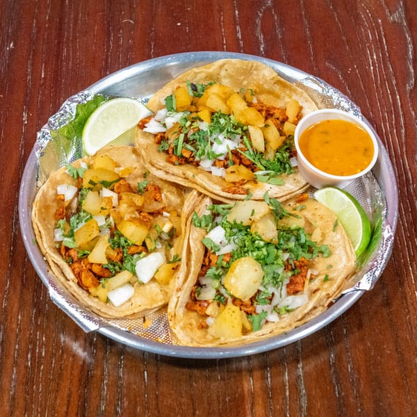 Three tacos with meat, potatoes, onions, cilantro, lime, and a side of sauce on a metal plate.