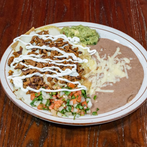 A plate of chilaquiles with meat, sour cream, guacamole, pico de gallo, and refried beans.