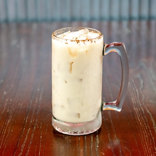 A tall glass mug filled with a creamy, light-colored drink, ice, and a cinnamon-sugar rim.